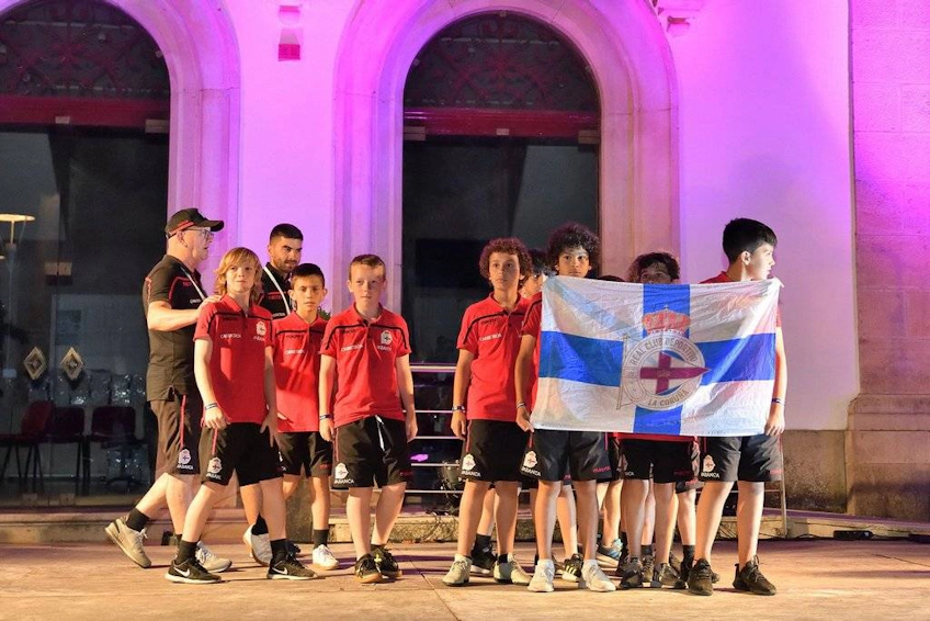 Gruppe av unge fotballspillere i røde uniformer med en trener, holder et flagg, på en nattlig sportsbegivenhet.