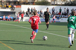 Ung spiller nummer 7 i rødt kontrollerer ballen med en dommer og tilskuere i bakgrunnen på Bahia de Roses Cup.
