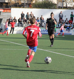 Joven jugador número 7 en rojo controlando el balón con un árbitro y espectadores al fondo en el torneo Bahia de Roses Cup.