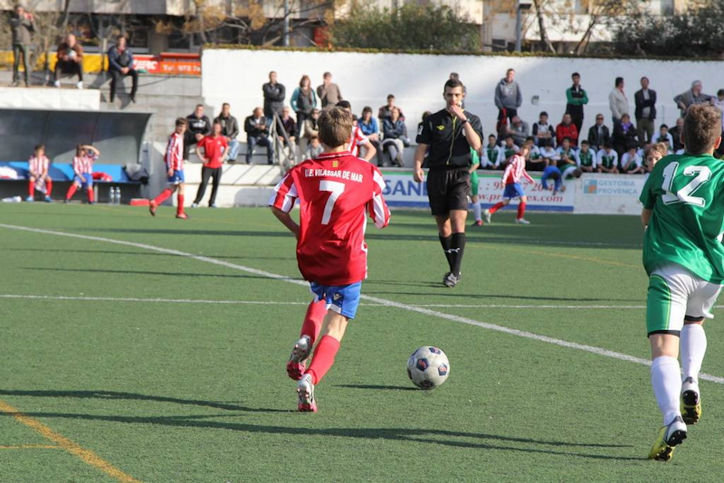 Kırmızı formayla genç oyuncu numara 7, Bahia de Roses Cup turnuvasında topu kontrol ediyor, arka planda hakem ve seyirciler var.