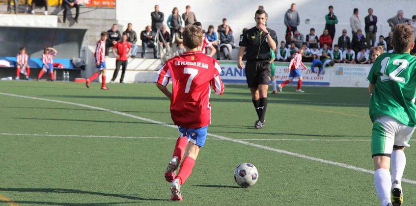 Jonge speler nummer 7 in het rood beheerst de bal met een scheidsrechter en toeschouwers op de achtergrond bij de Bahia de Roses Cup.