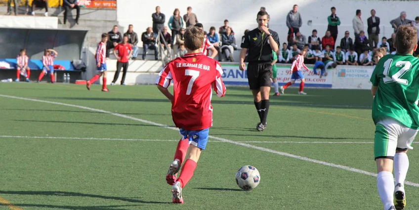 Joven jugador número 7 en rojo controlando el balón con un árbitro y espectadores al fondo en el torneo Bahia de Roses Cup.