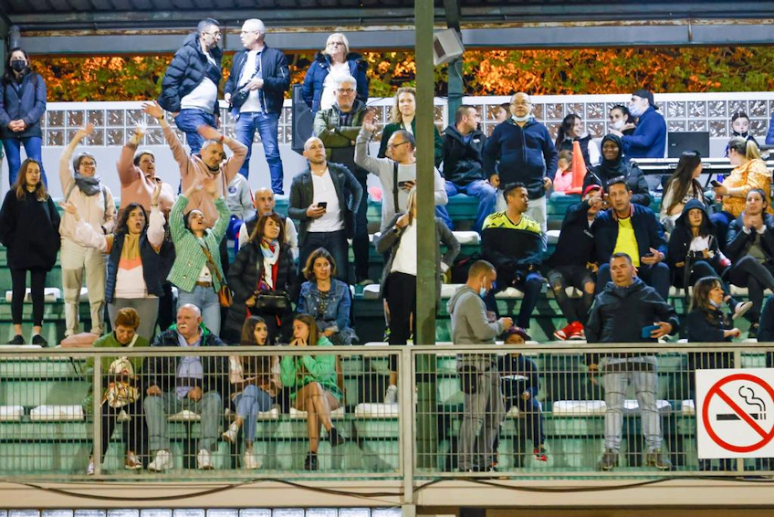 Spettatori con espressioni diverse che godono di una partita di calcio alla Coppa Bahia de Roses, cartello di divieto di fumo visibile.