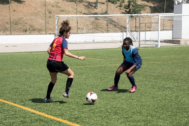 Female soccer player in red and blue stripes dribbling against an opponent in dark blue on a sunny day.