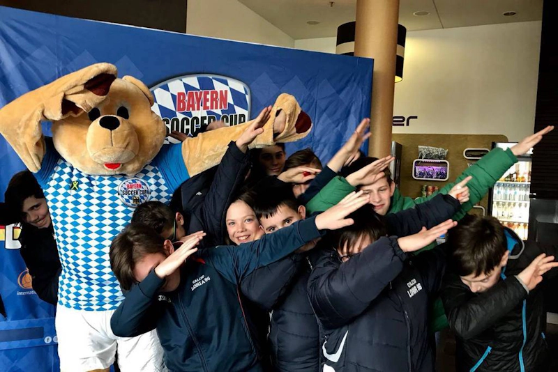 Grupo de niños con un gran oso de peluche frente al logo de Bayern Soccer Cup