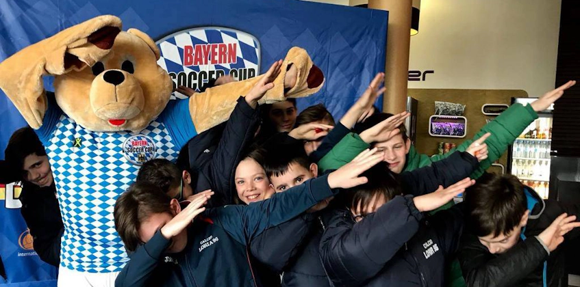 Grupa dzieci z dużym pluszowym misiem przed logo Bayern Soccer Cup