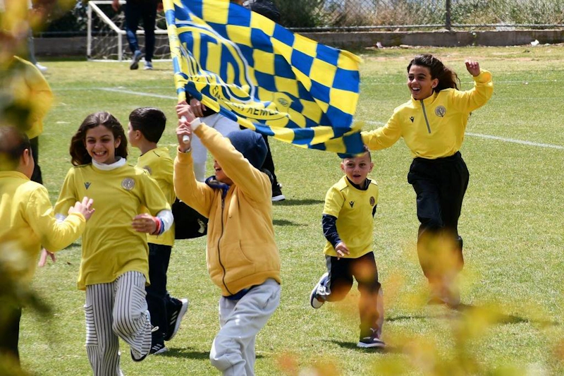पीली शर्ट में खुश बच्चे झंडे के साथ फुटबॉल मैदान पर दौड़ते हुए