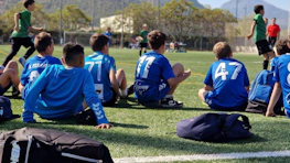 Pojkar i blå tröjor sitter vid sidlinjen och tittar på en fotbollsmatch under Esei Madrid Elite Cup-turneringen.