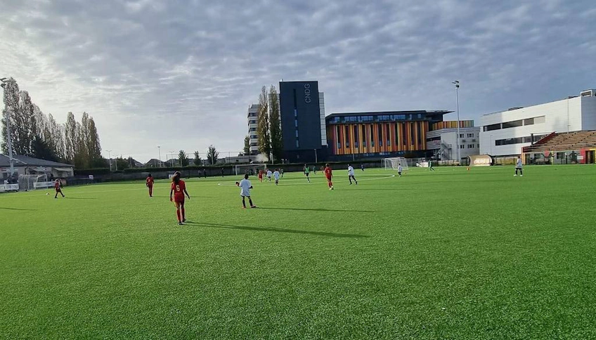 Jogadores no torneio de futebol Scimemi Cup em campo verde com edifícios ao fundo