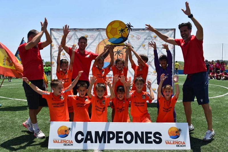 Ifjúsági labdarúgócsapat ünnepli a győzelmet a Valencia Beach Tornán edzőkkel és 'Campeones' zászlóval.