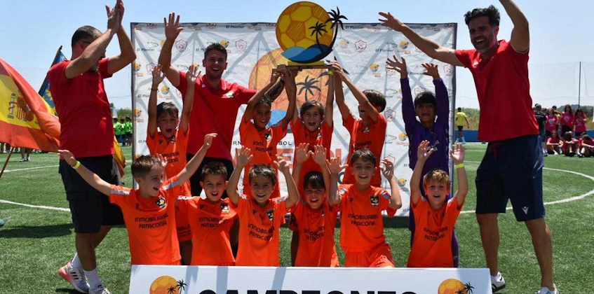Equipo juvenil de fútbol celebra victoria en Torneo Valencia Beach con entrenadores y pancarta 'Campeones'.