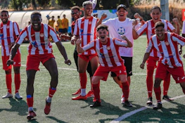 Ομάδα ποδοσφαιριστών με κόκκινα και λευκά ριγέ στολές πανηγυρίζει νίκη στο γήπεδο
