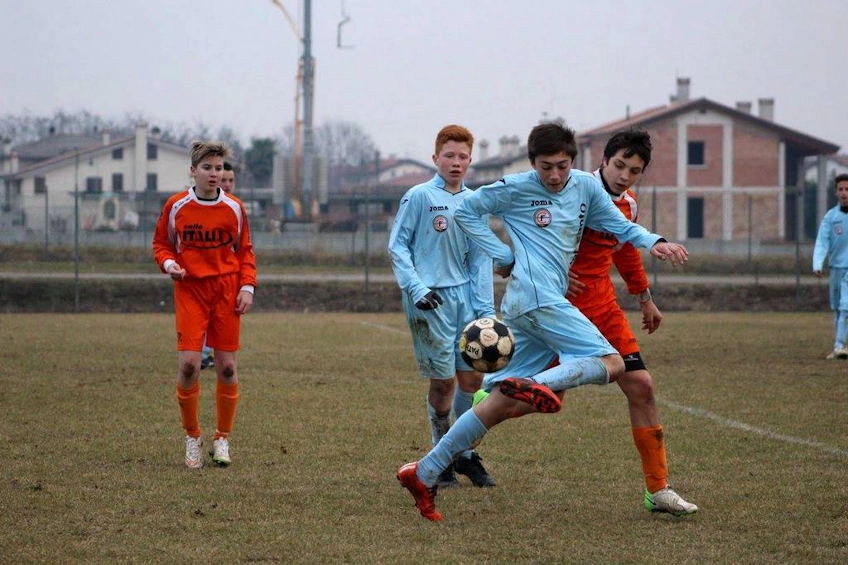 Teini-ikäiset jalkapalloilijat toiminnassa pilvisenä päivänä, yksi vaaleansinisessä pitelee palloa.