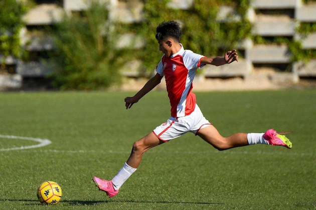 Młody gracz w czerwono-białym stroju wykonujący potężne uderzenie podczas meczu piłkarskiego w słoneczny dzień.