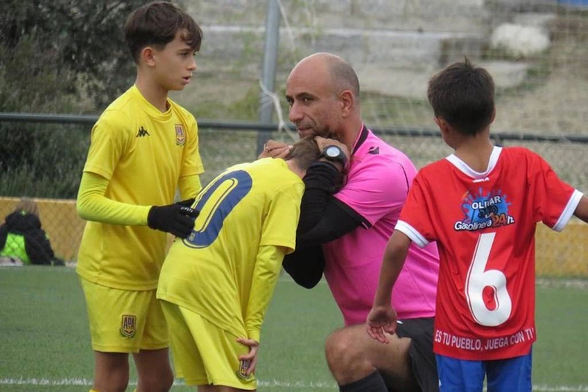 A youth football goalkeeper in yellow talking to a referee comforting a player in red