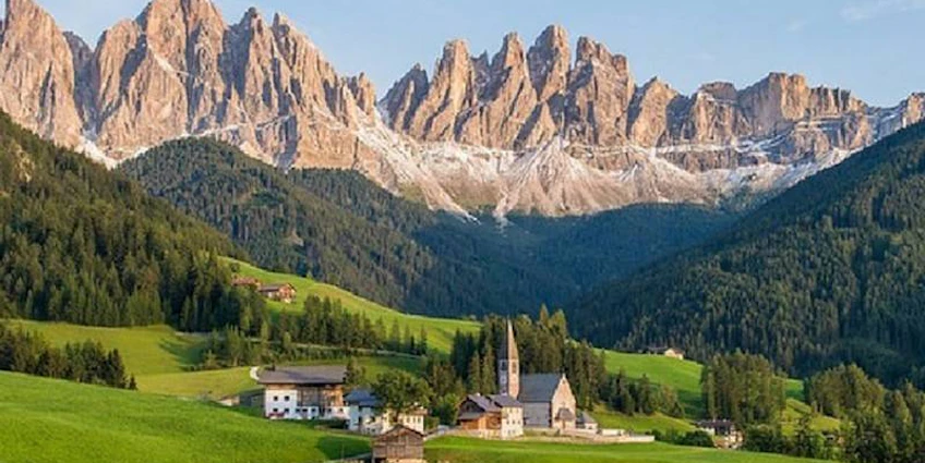 Aldeia nas Dolomitas com uma igreja no torneio Grand Prix Dolomites Summer Trophy, rodeada por montanhas e florestas.