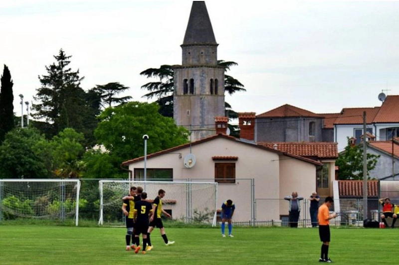 Partido de fútbol en el Grand Prix Poreč Summer Trophy con una iglesia y árboles de fondo.