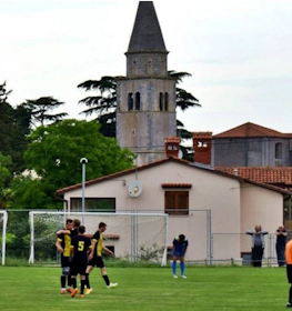 Fotballkamp på Grand Prix Poreč Summer Trophy med en kirke og trær i bakgrunnen.
