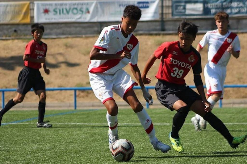 Meci de fotbal la turneul U18 Madrid Youth Cup Summer, jucători în uniforme roșii și albe se luptă pentru minge.