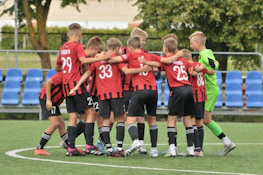 Football team in red and black uniforms at Summer Finest League tournament, players gathered in a circle.