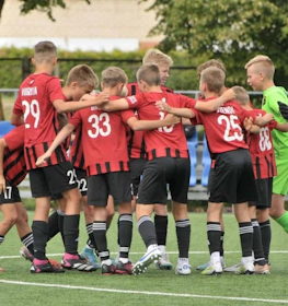Football team in red and black uniforms at Summer Finest League tournament, players gathered in a circle.