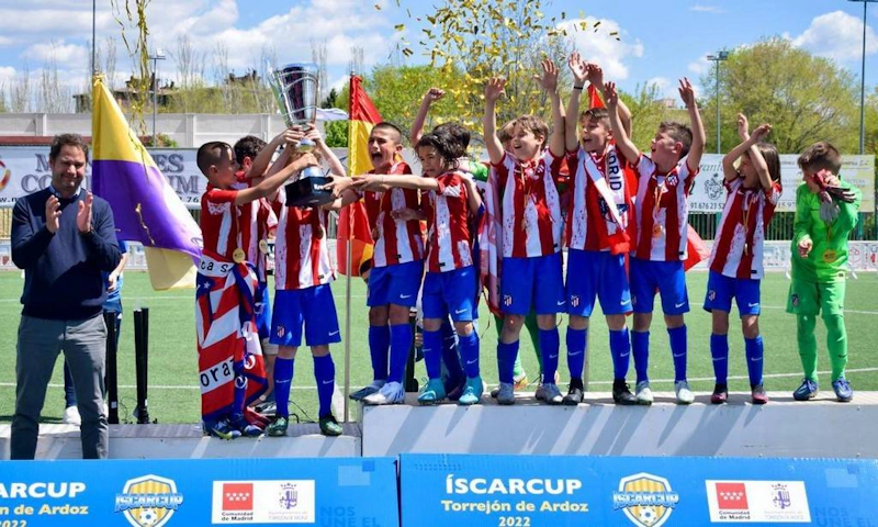 Young soccer players in red and white striped uniforms celebrate victory with a trophy at the ÍscarCup soccer tournament.