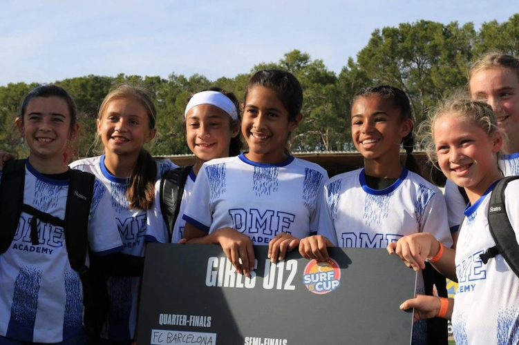 Группа молодых футболисток из академии DME держит знак четвертьфинала на турнире Surf Cup.